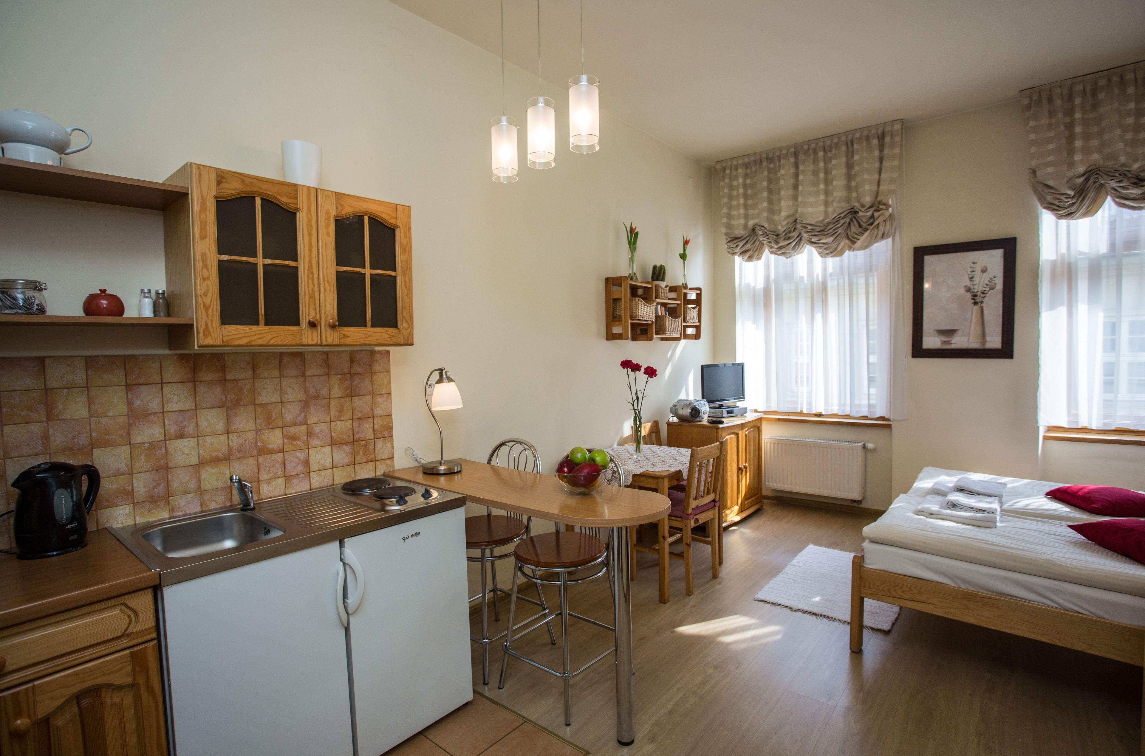 A compact kitchen with a sink, stove, and wooden cabinets. There is a dining area with a table and chairs, and a bed with a red pillow.