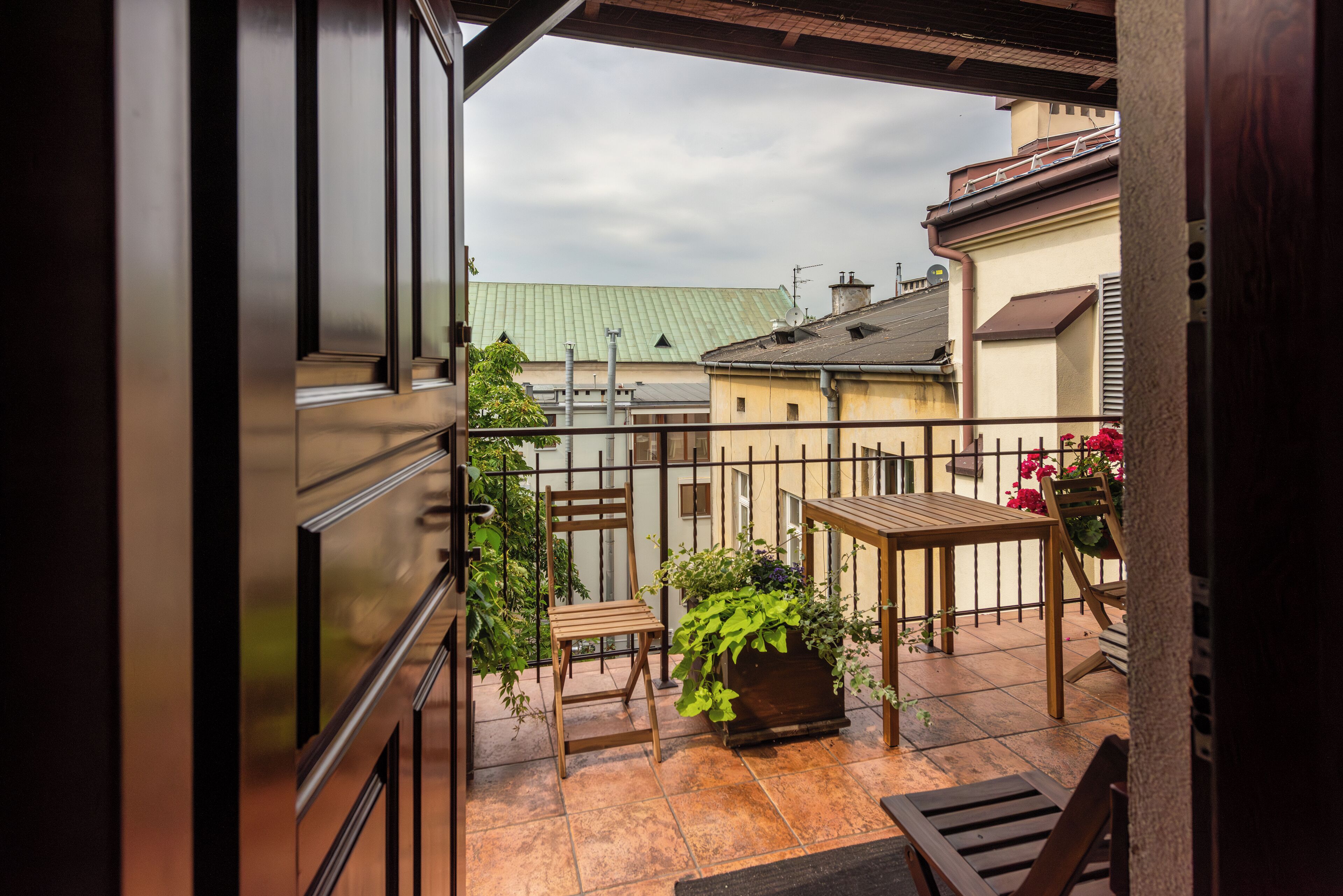 Varanda com mesa e cadeiras de madeira, plantas em vasos e vista para edifícios vizinhos.
