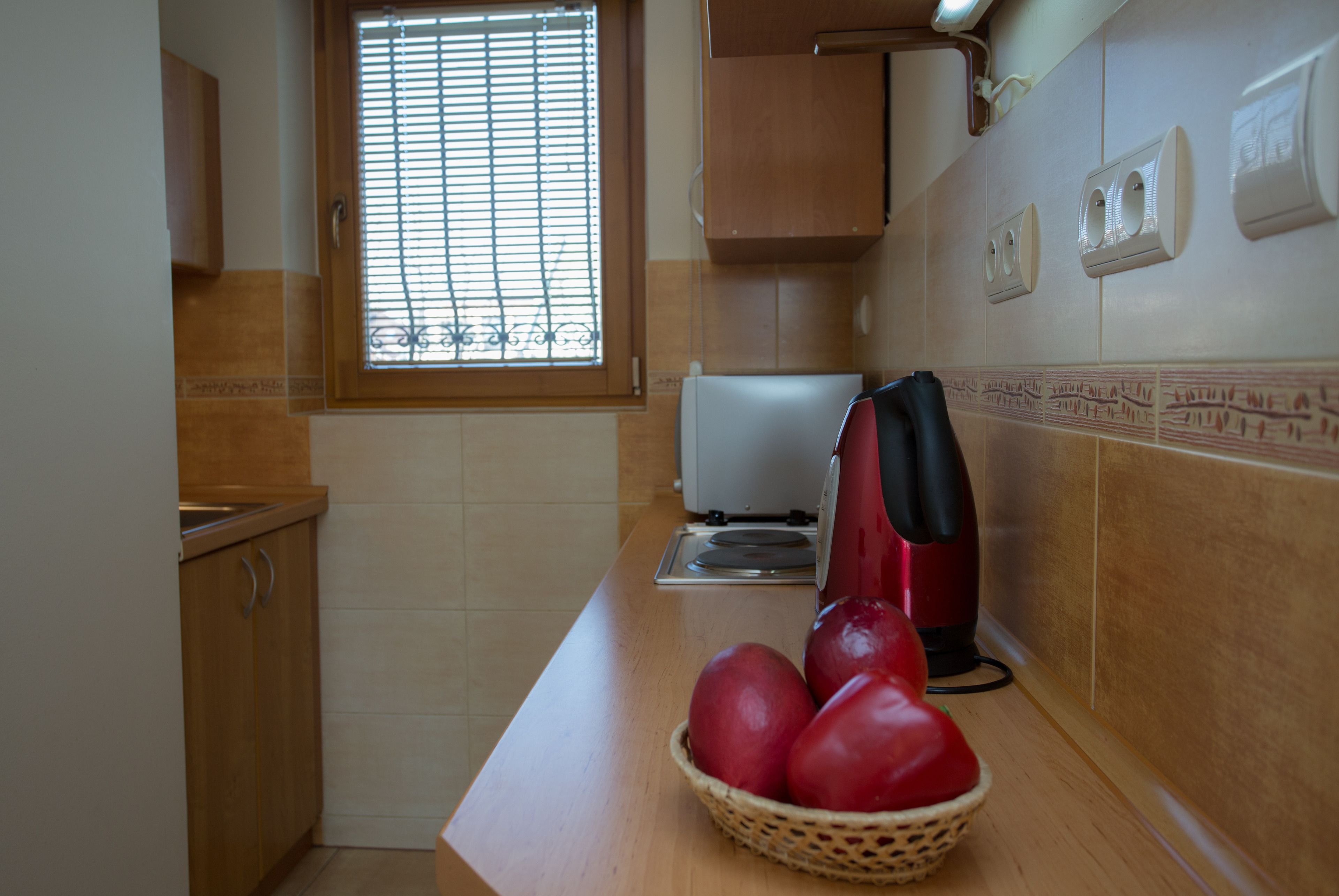 Une cuisine avec des meubles de rangement en bois, une fenêtre avec des stores, une bouilloire rouge et un panier de pommes rouges posé sur le comptoir.