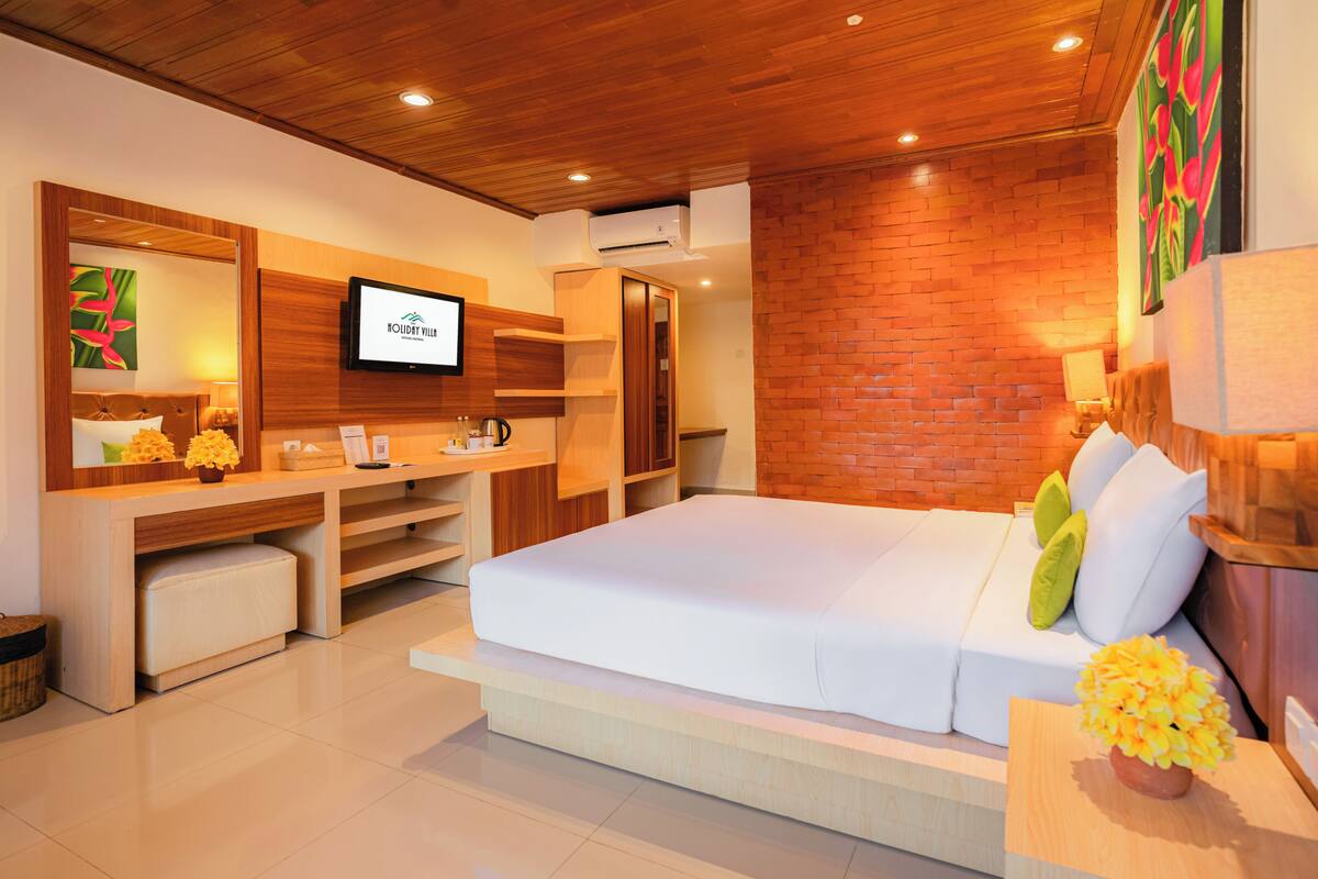 Quarto de hotel moderno com teto de madeira, televisão de tela plana, cama com roupa de cama branca, escrivaninha com cadeira e parede decorativa de tijolos.