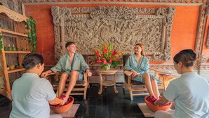 A room with a traditional carved wall, a round wooden table with a vase of flowers, and four people seated on the floor. - Wina Holiday Villa (Kuta)