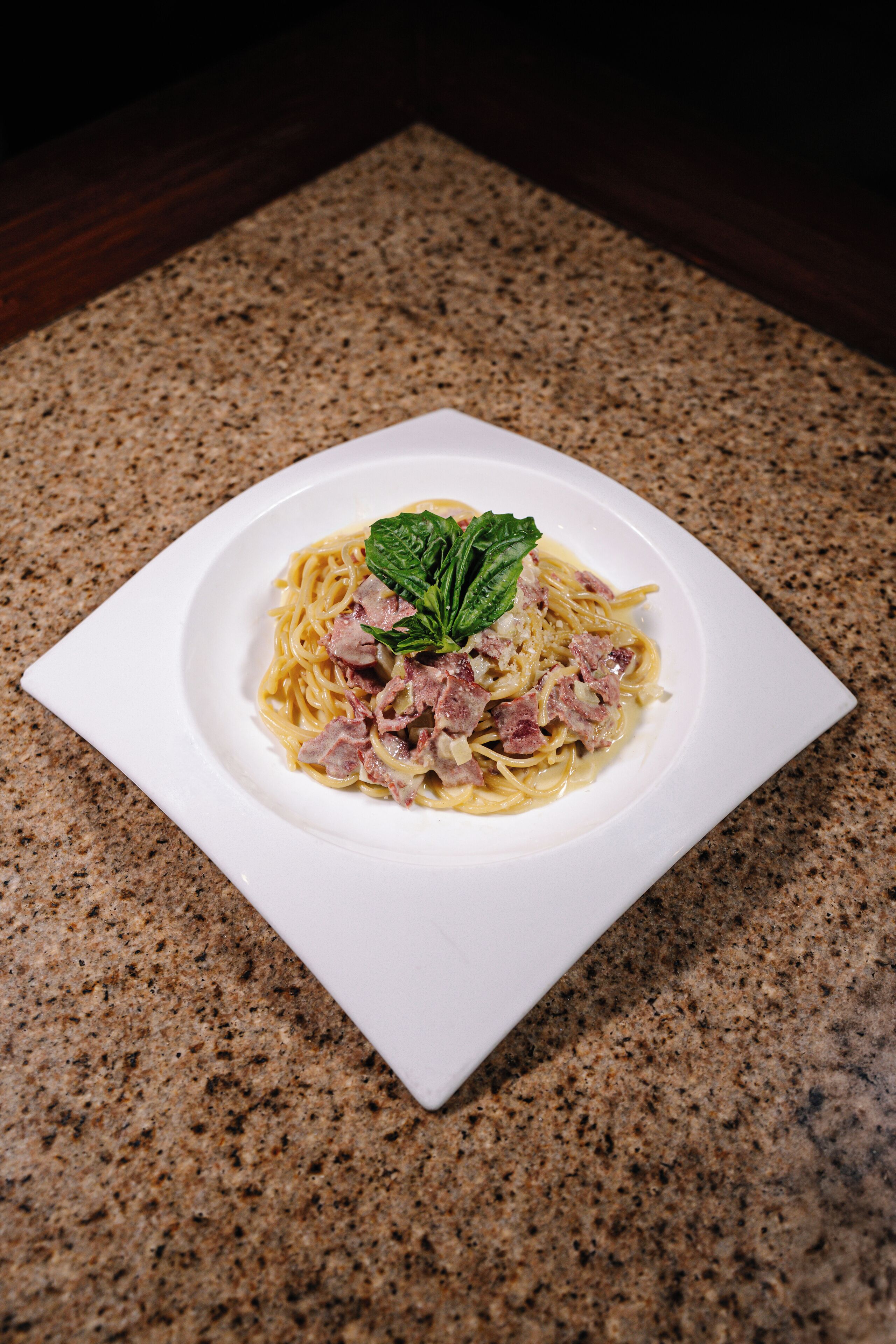 Un plato blanco cuadrado con pasta y carne sobre una encimera de granito moteado.