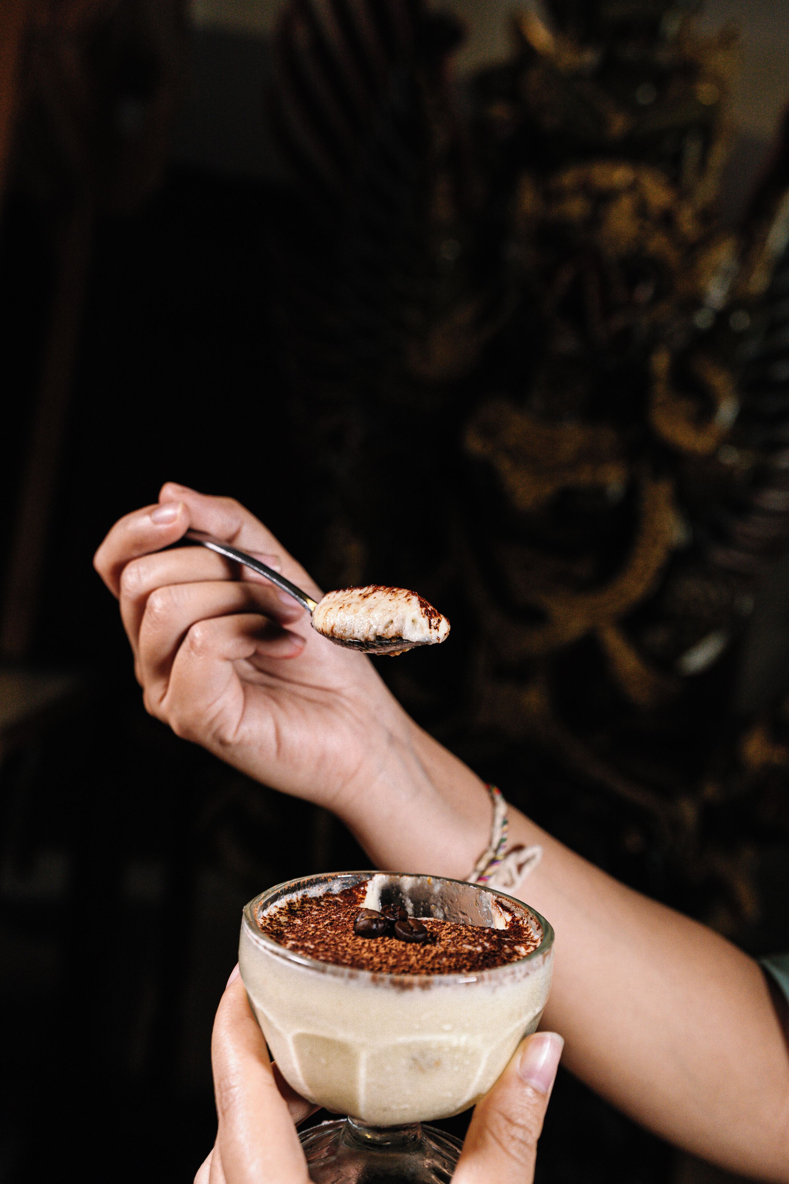A hand holding a glass with a dessert, possibly a tiramisu, with a spoon lifting a piece.