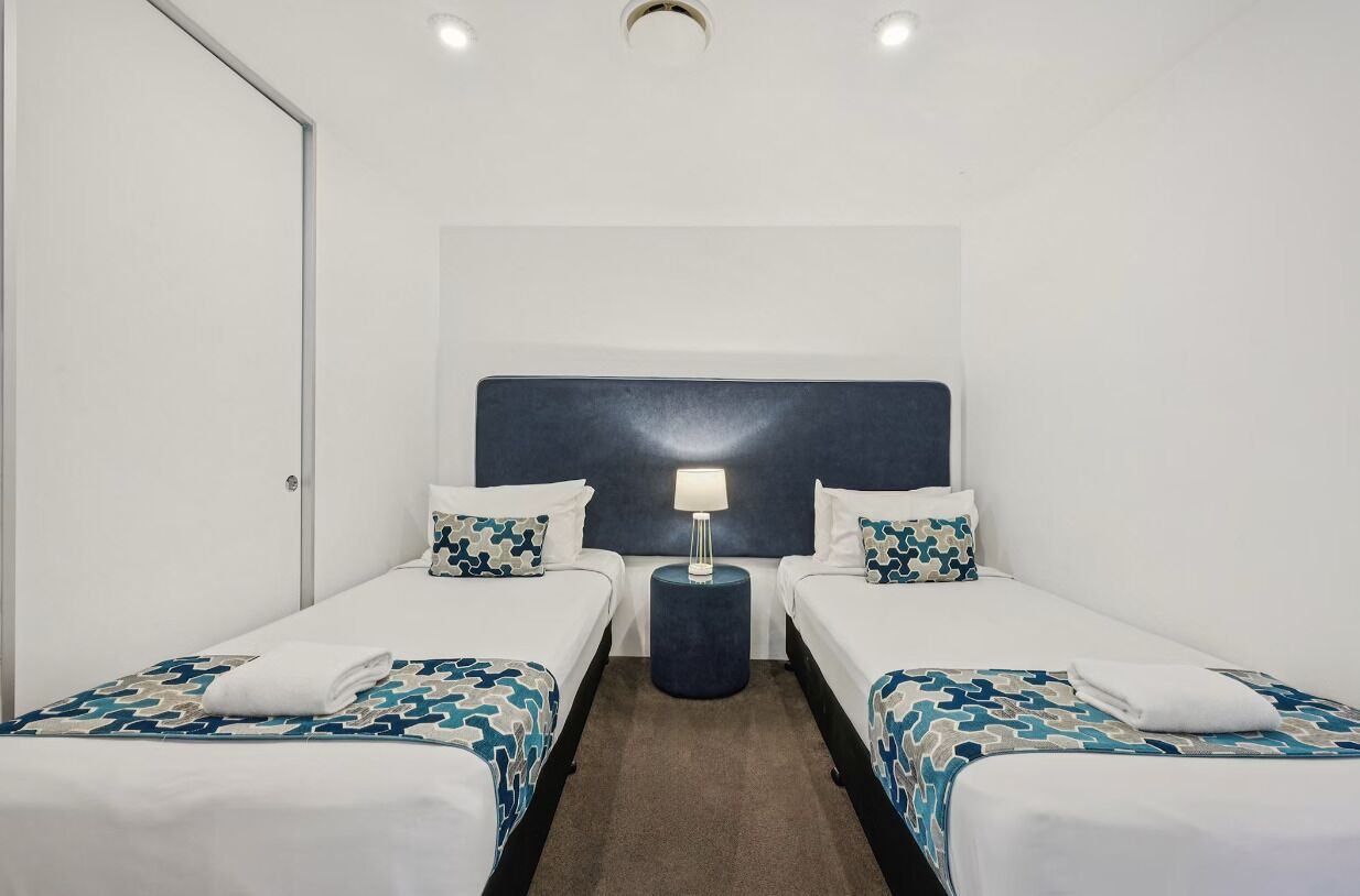 Habitación de hotel con dos camas individuales, cabecera, mesita de noche con lámpara y piso alfombrado.