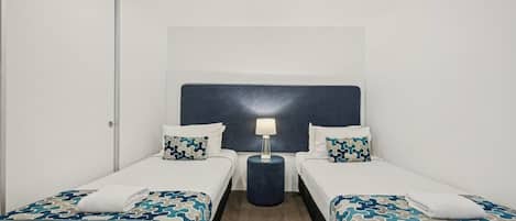 Habitación de hotel con dos camas individuales, cabecera, mesita de noche con lámpara y piso alfombrado.