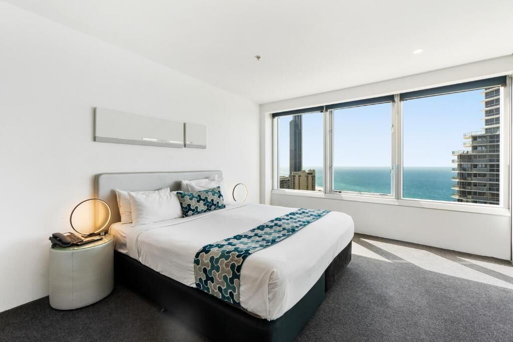 A modern hotel room with a large bed, a bedside table with a lamp, a phone, and a speaker. There is a large window offering a view of the ocean and buildings.