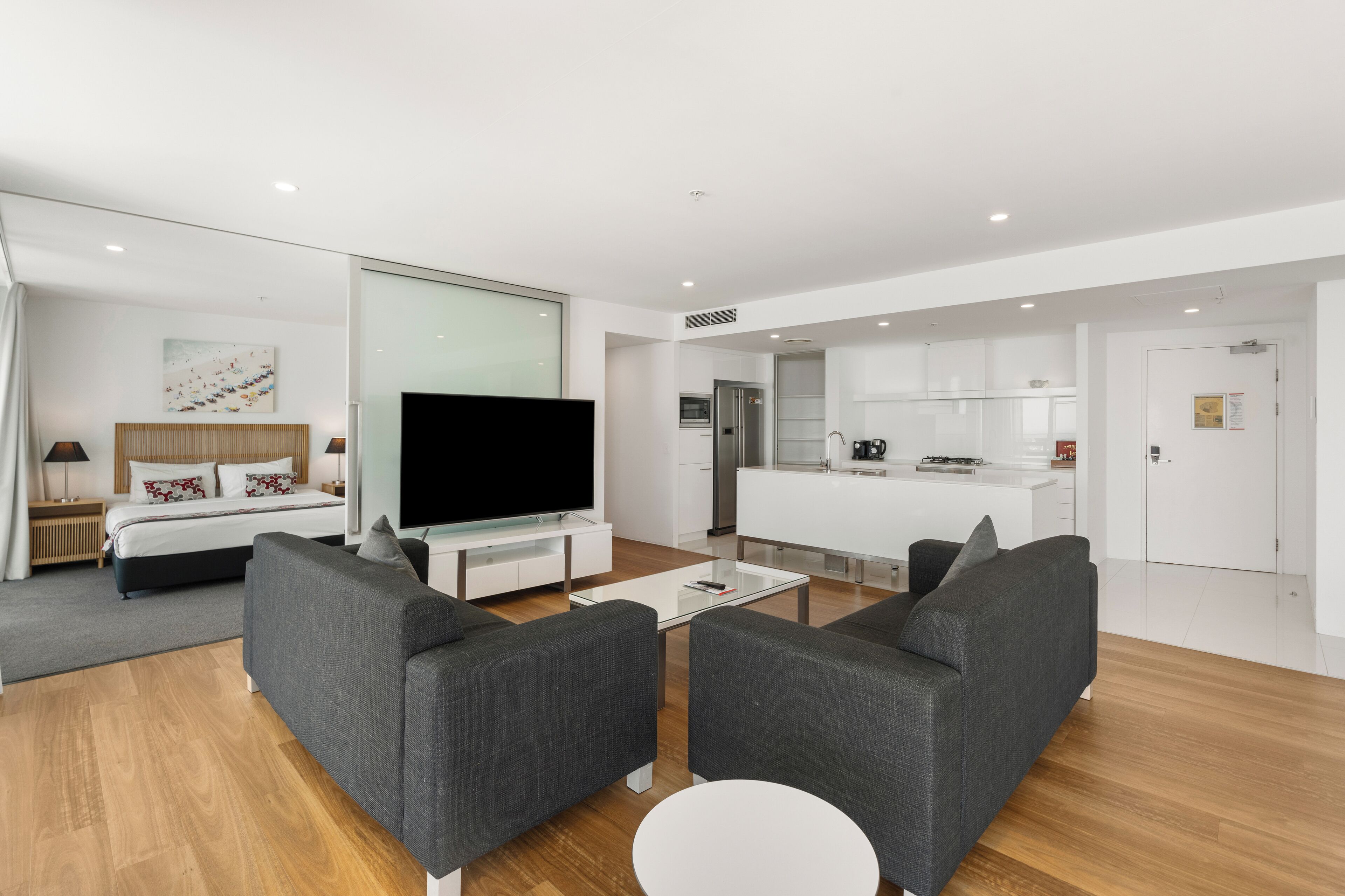 A modern living room with a flat-screen TV, grey sofas, a coffee table, and a kitchen area in the background.