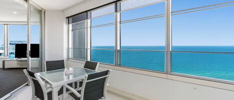 A modern room with a glass table and chairs, large windows with a view of the sea, and a sliding glass door leading to another area.