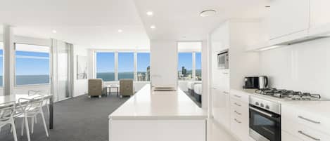 Une cuisine moderne avec des meubles blancs, un îlot central doté d’un évier et une vue sur l’océan grâce à de grandes fenêtres.