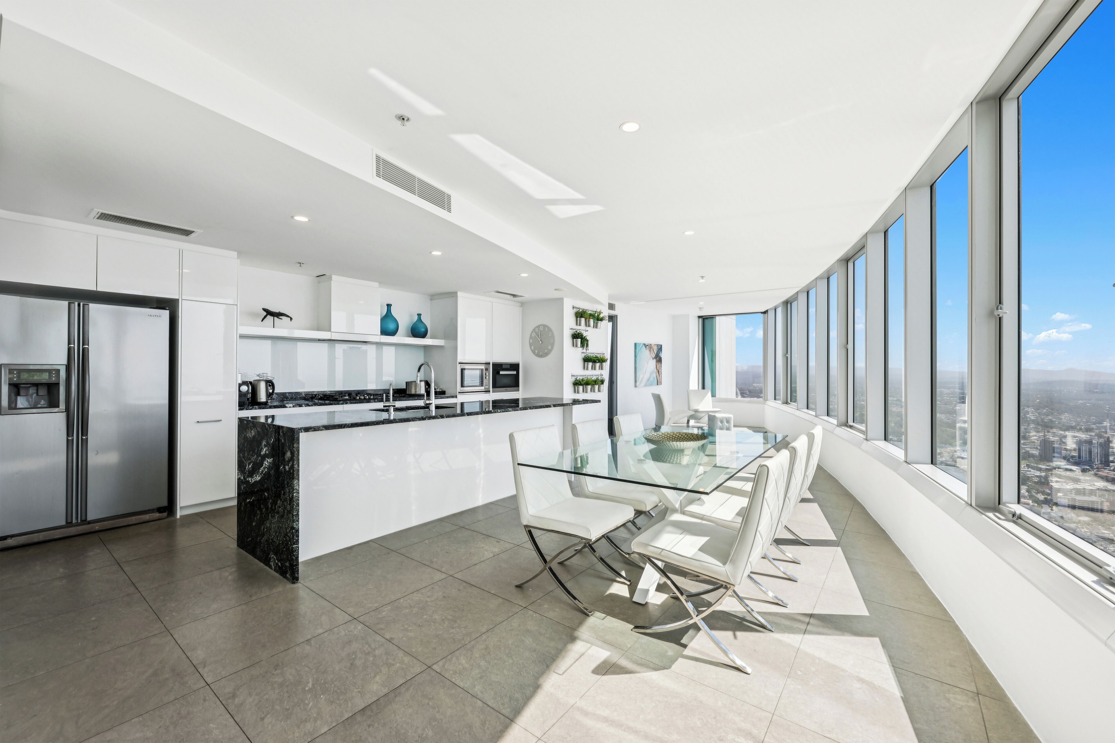 Una cocina moderna con una mesa de comedor y sillas de vidrio, un refrigerador de acero inoxidable y vistas a la ciudad a través de amplios ventanales.