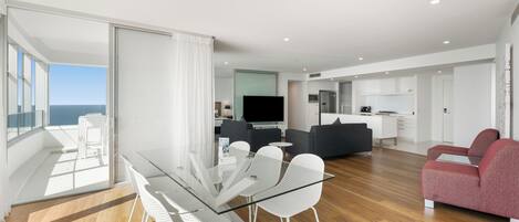 A modern living room with a glass dining table, white chairs, a red sofa, and a kitchen area in the background.