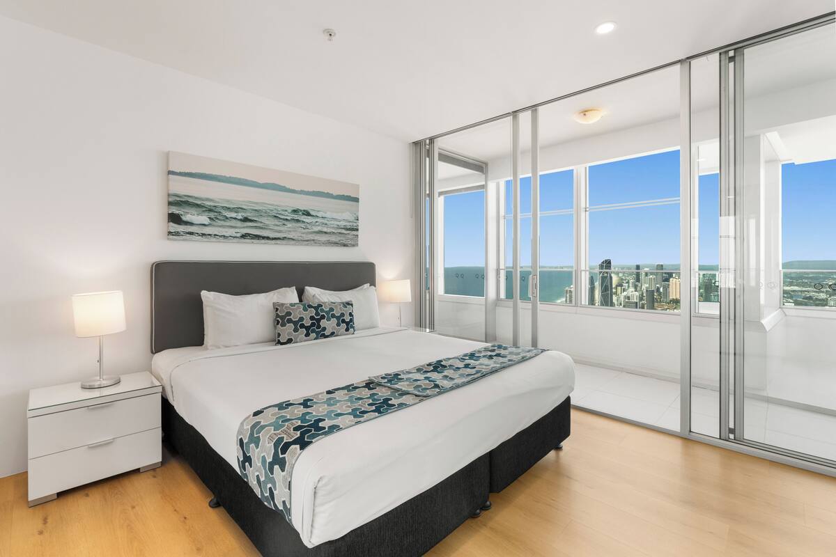 A modern bedroom with a large bed, a city view through floor-to-ceiling windows, and a cityscape visible outside.