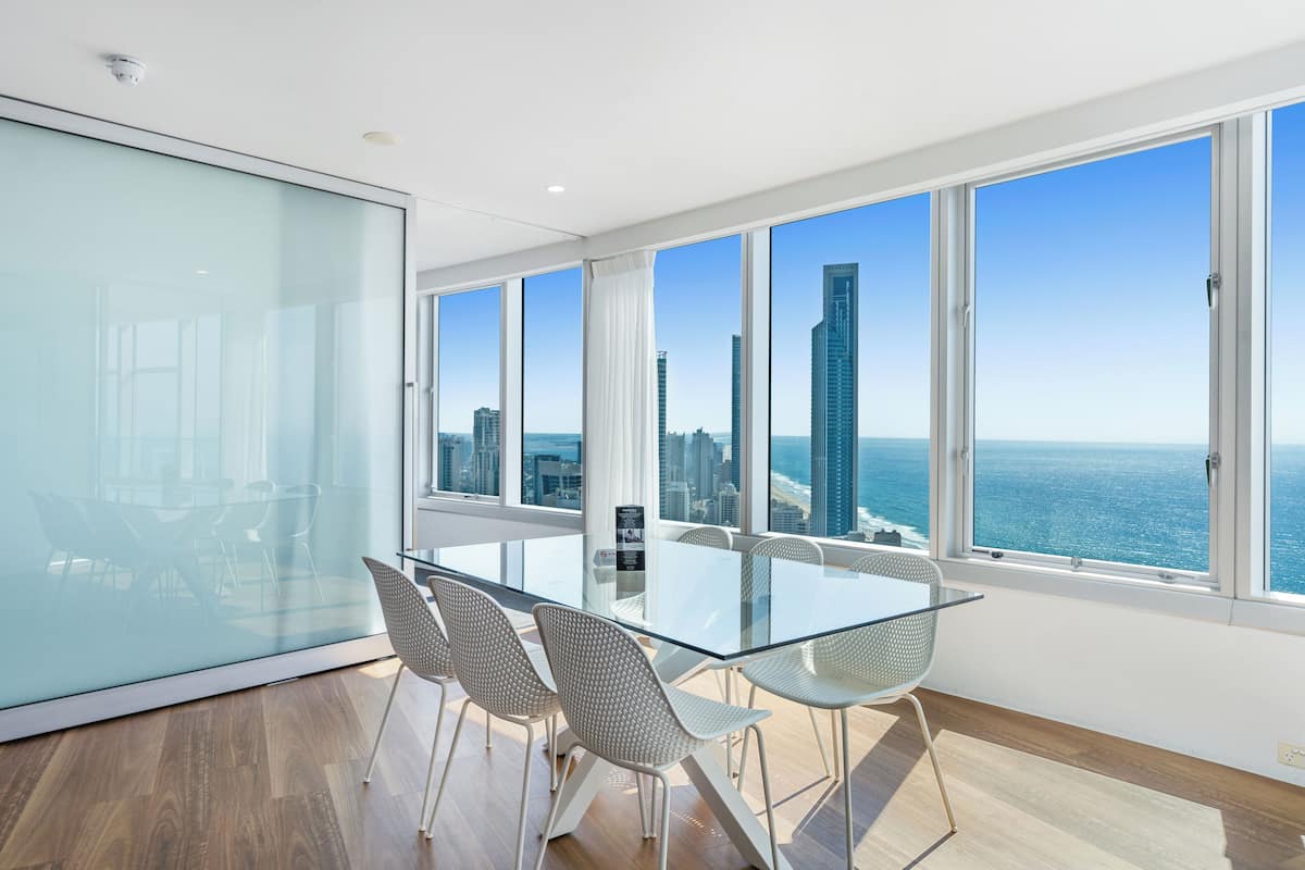 A modern meeting room with a glass table, white chairs, and a view of the ocean and city skyline through large windows.