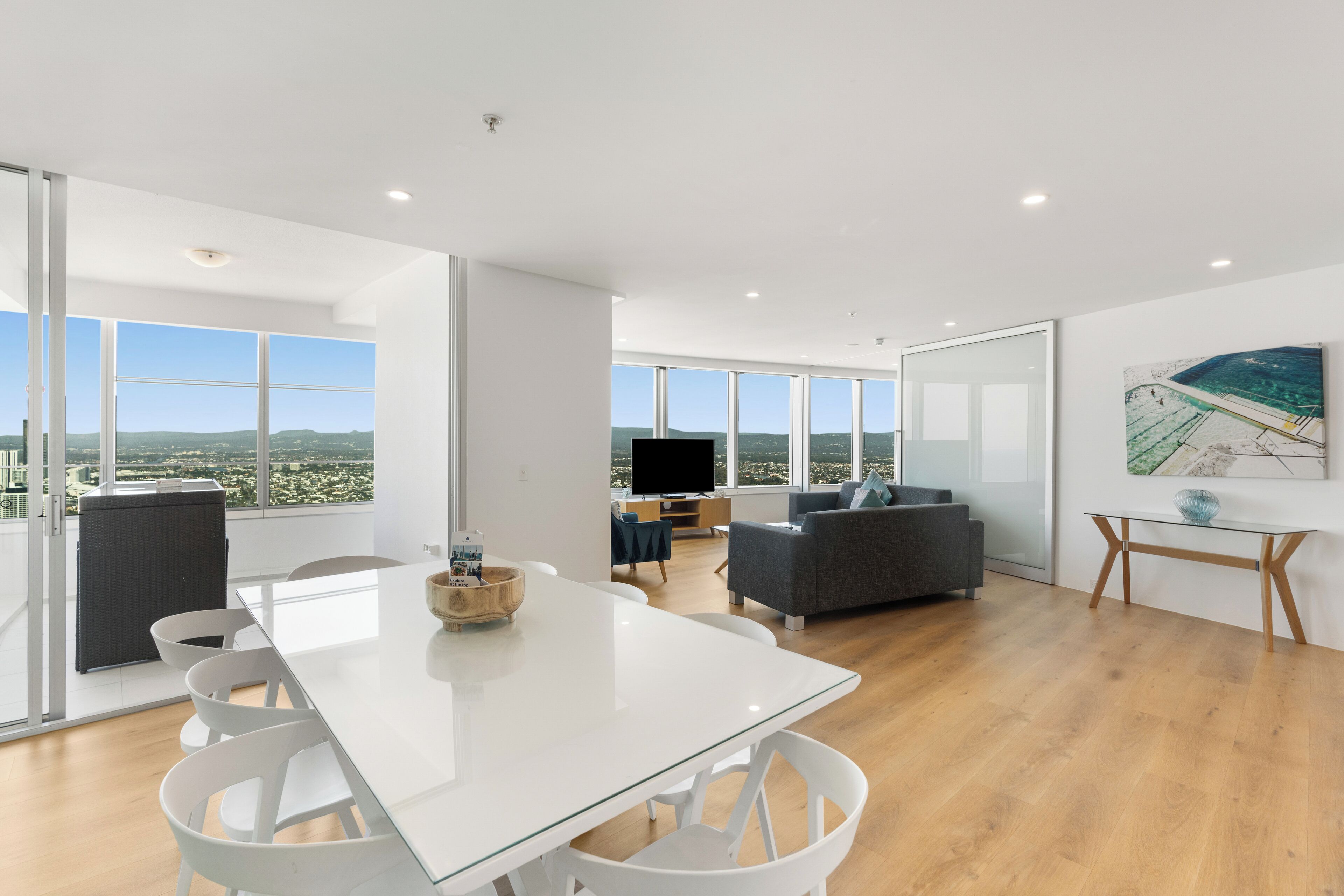 Una sala de estar moderna con una mesa de comedor de cristal, sillas blancas, un sofá gris y una televisión.