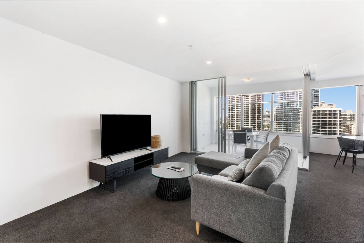 A modern living room with a flat-screen TV, a grey sofa, a glass coffee table, and a dining area with a glass partition.