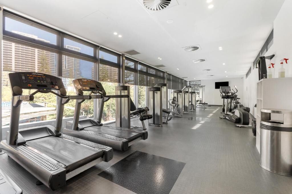A modern gym with treadmills, weight machines, and a large window providing a view of buildings outside.