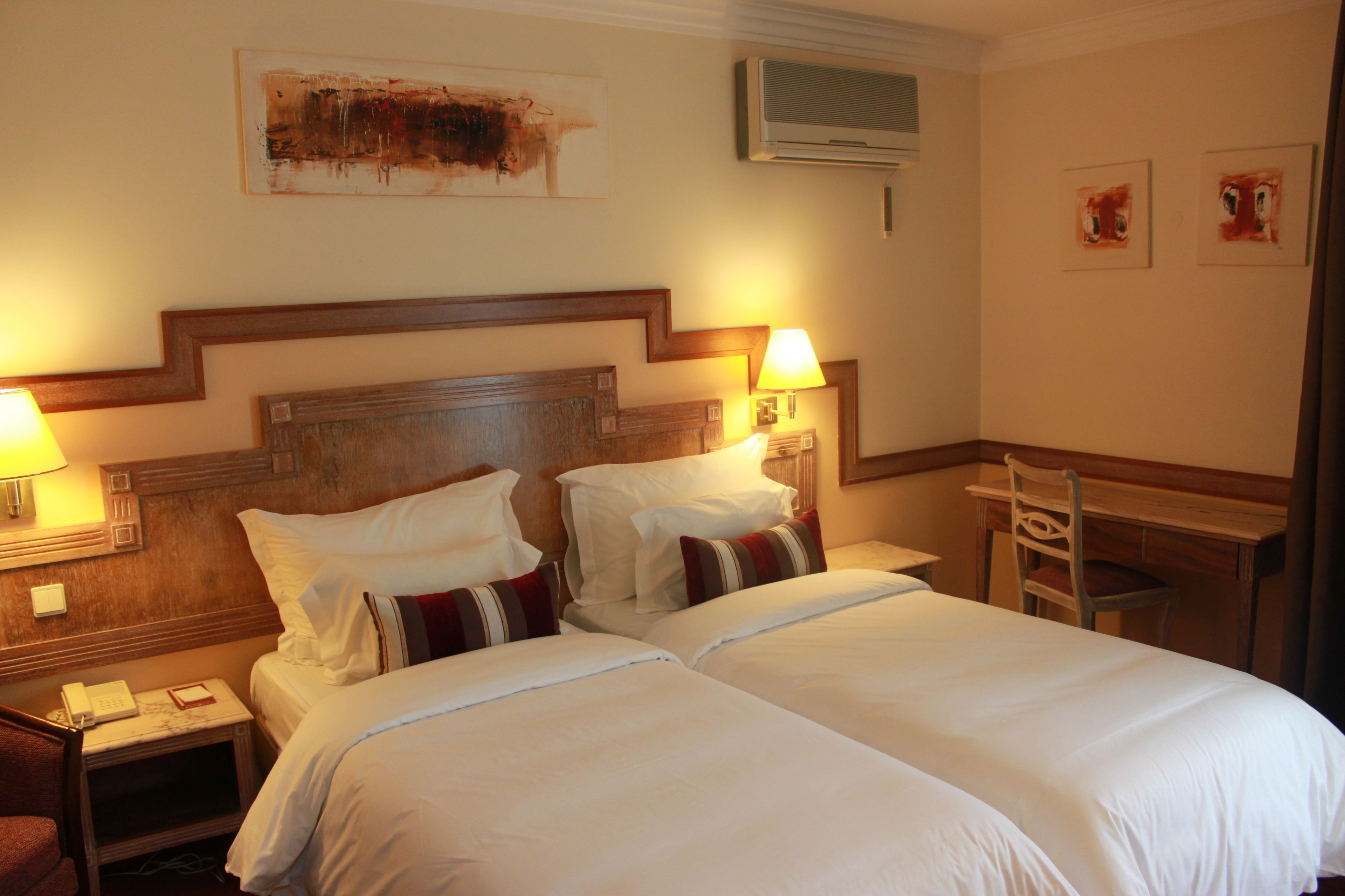 Ein Hotelzimmer mit zwei Betten, einem Holz-Kopfteil, Nachttischlampen, einem Schreibtisch und einem Stuhl.