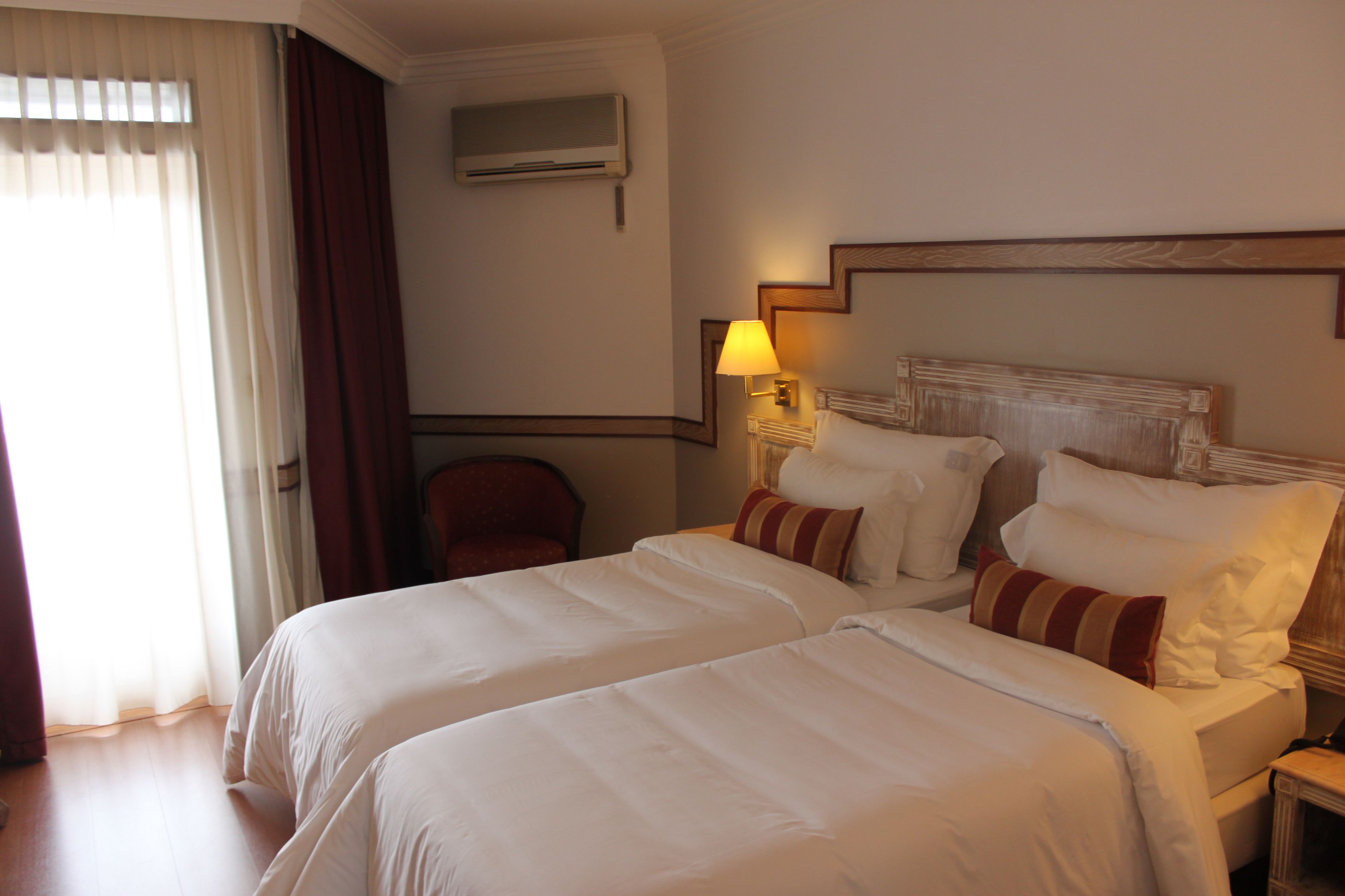 A hotel room with two beds, a chair, a lamp, and an air conditioning unit.