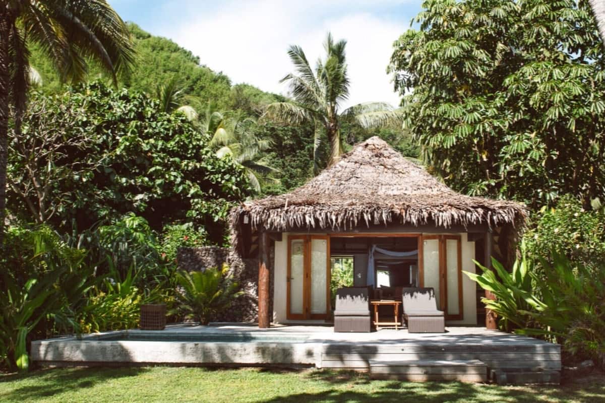 A thatched-roof hut with a wooden door and windows, surrounded by lush greenery and palm trees.