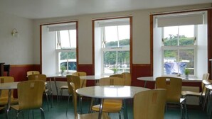 A dining area with round tables and yellow chairs, large windows with white frames, and a view of greenery outside. - Tobermory Youth Hostel (Isle of Mull)