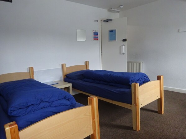 A room with two wooden beds with blue bedding, a small bedside table, a whiteboard, and a door with a security camera.