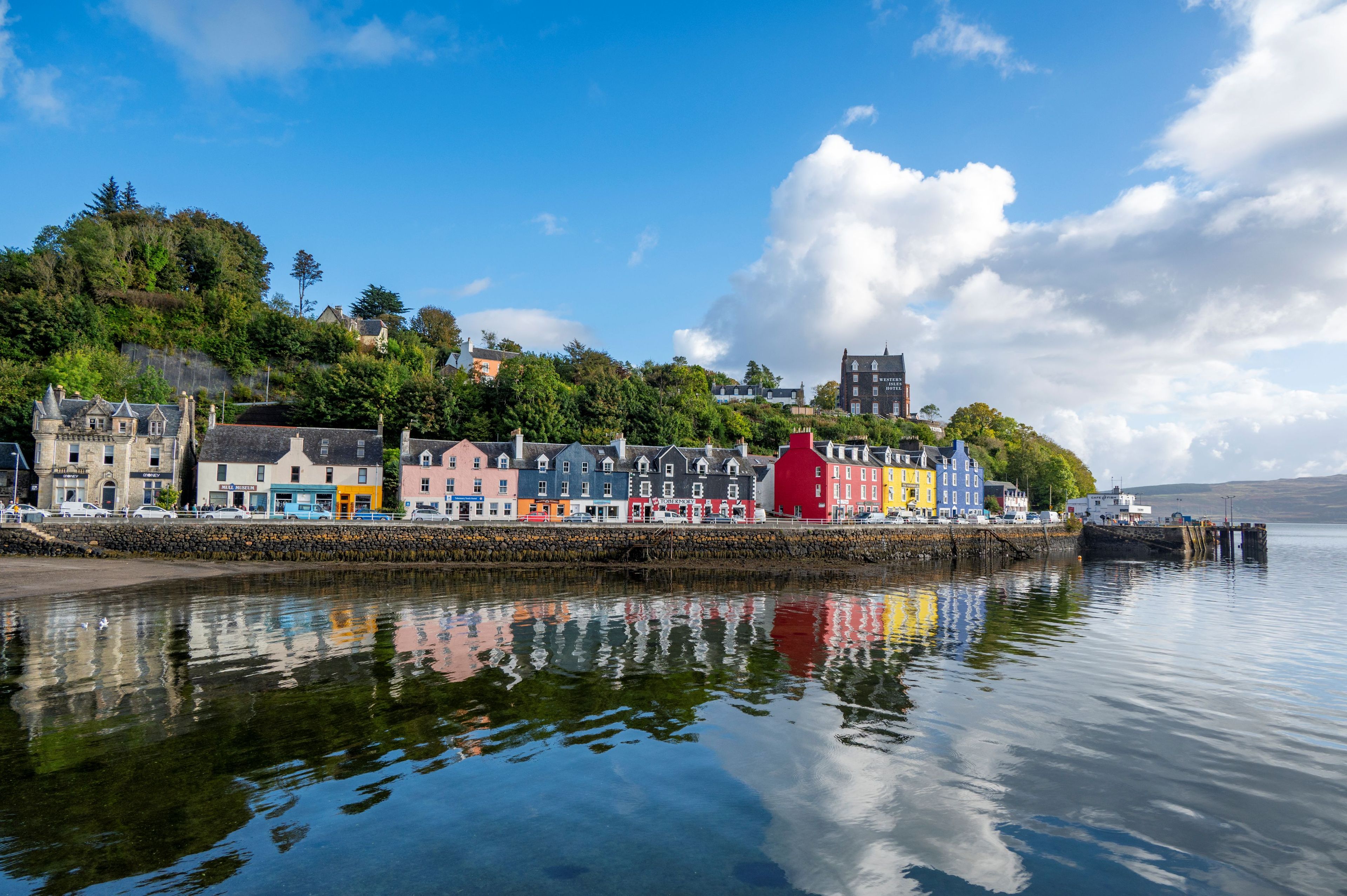 A waterfront town with colorful buildings, a stone wall, and a calm body of water reflecting the sky and structures.
