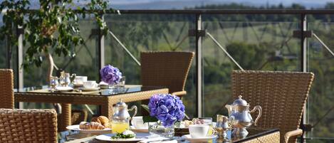 A wicker dining table set with breakfast items, a vase of flowers, and a scenic view.