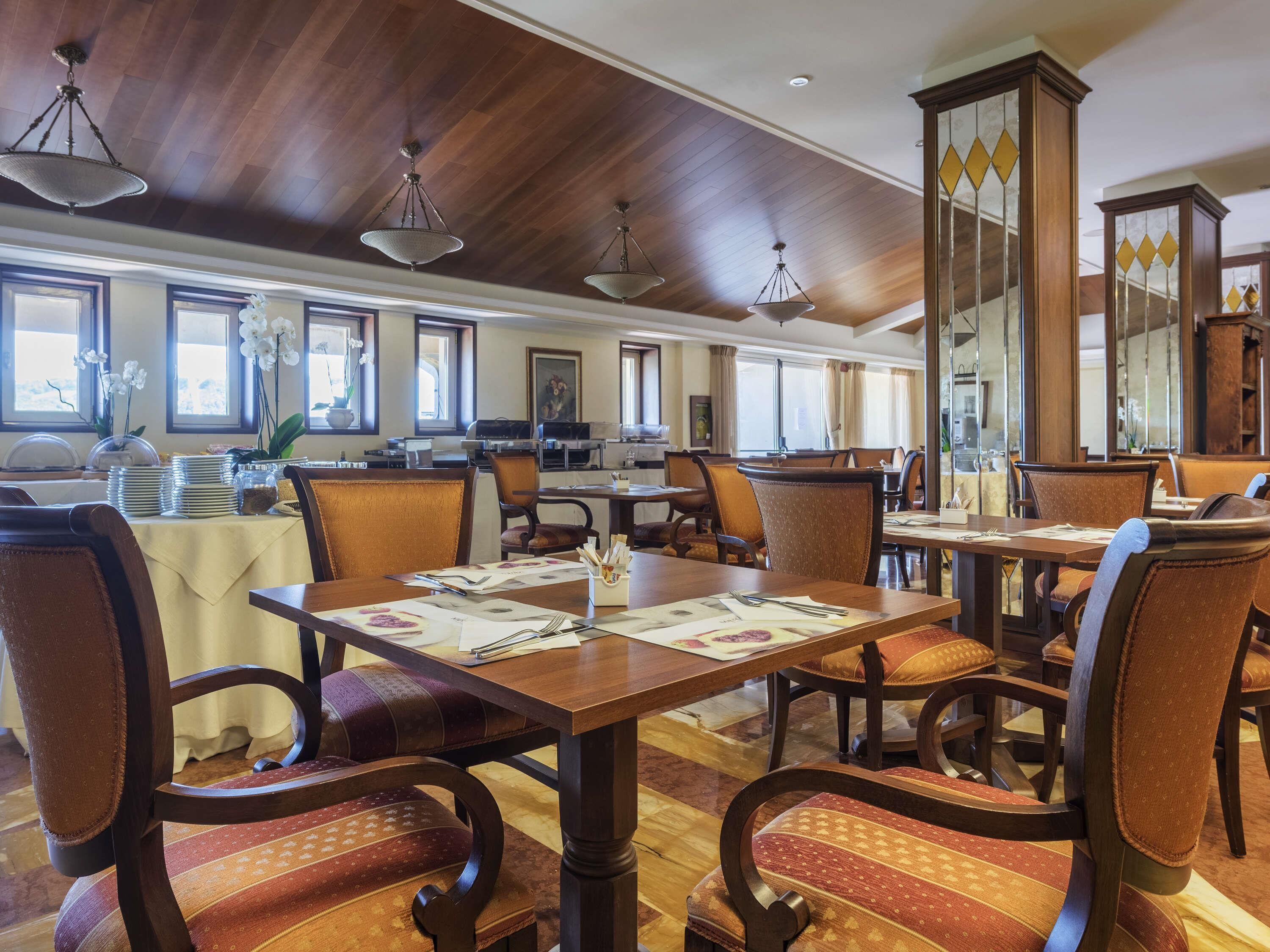 A spacious dining area with wooden tables and chairs, large windows, and a high ceiling with hanging lights.