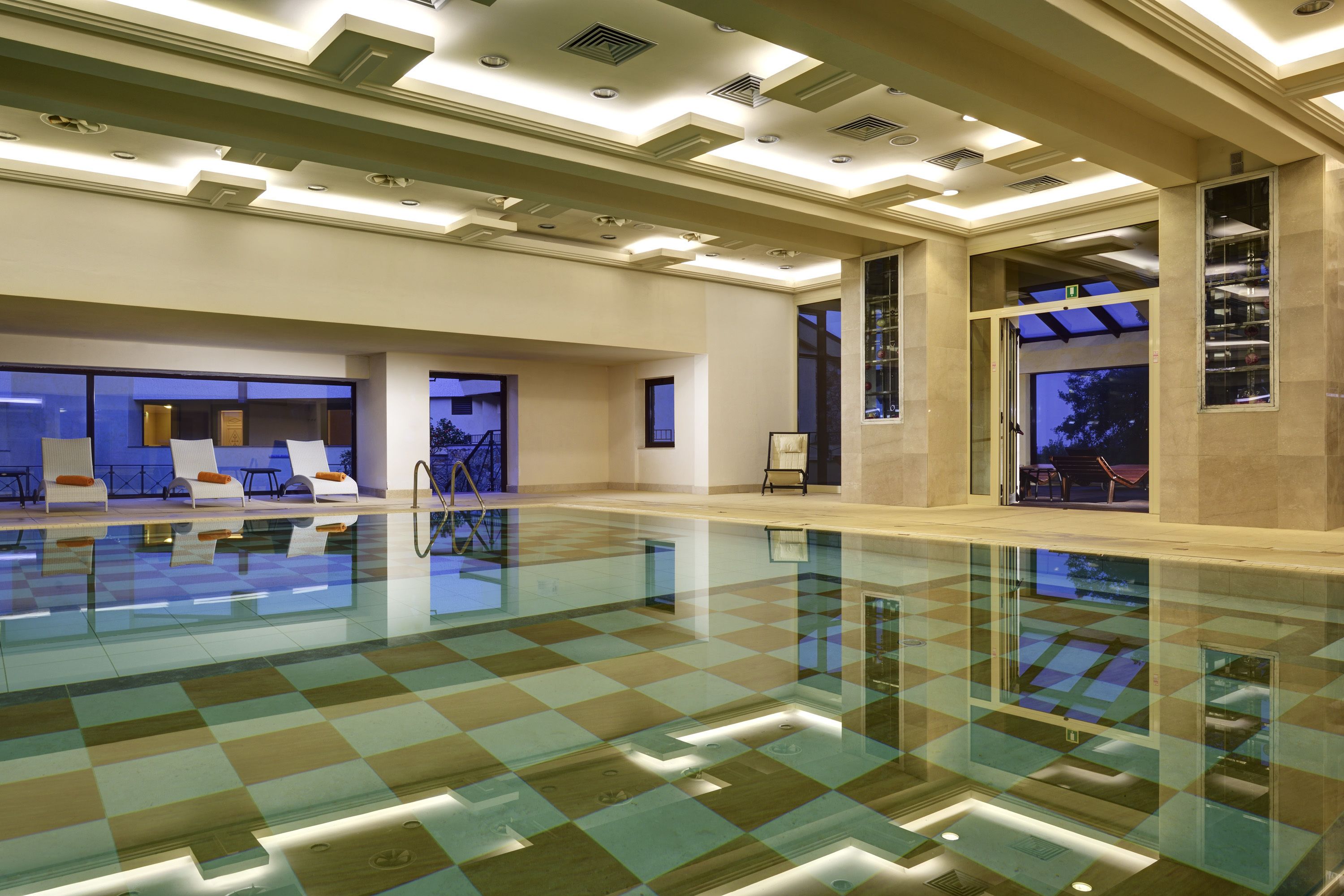 A spacious indoor swimming pool with a checkered floor, recessed lighting, and a view of the outdoors through large windows.