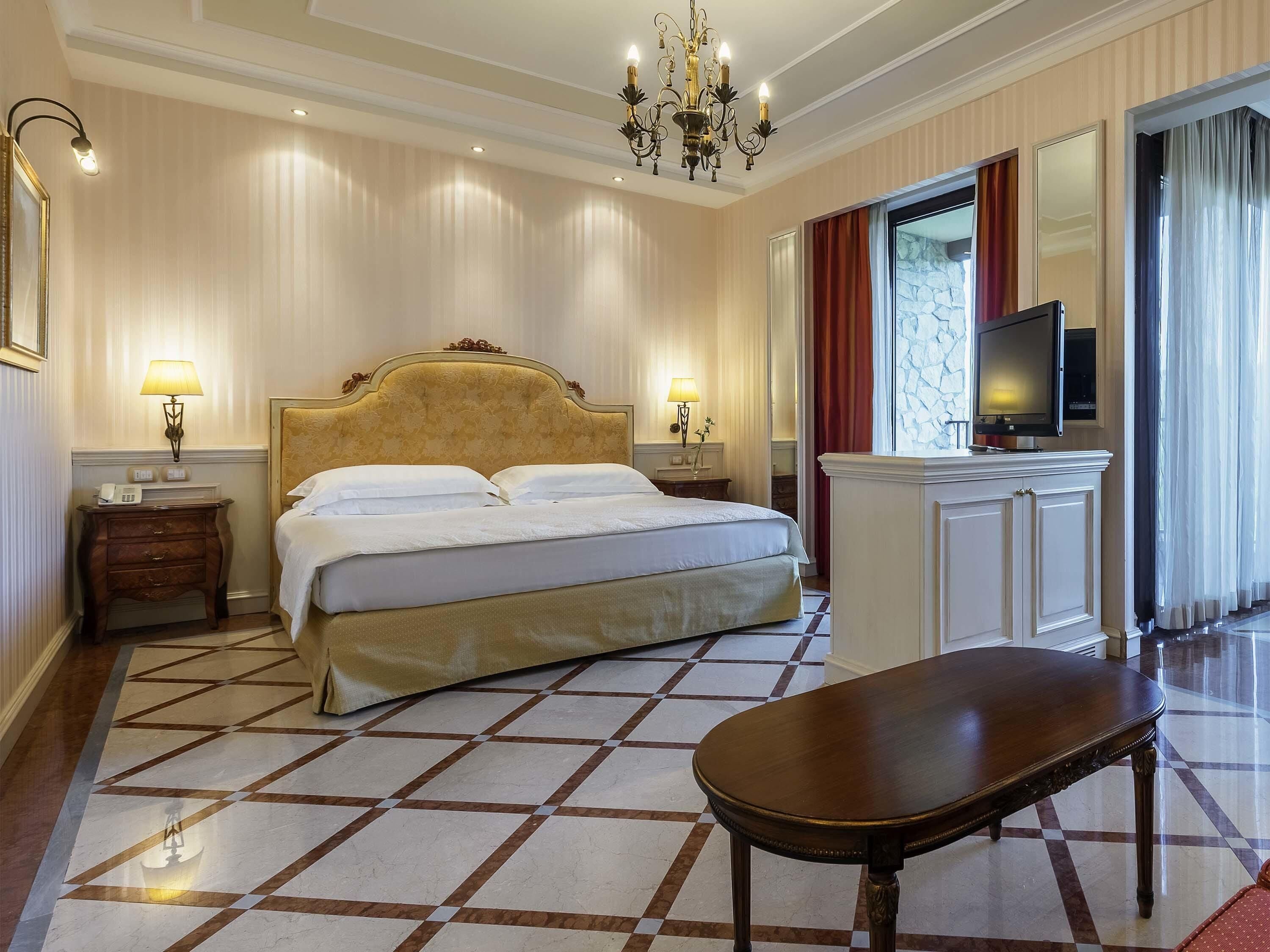 Una habitación de hotel con una cama grande, lámparas de noche, un televisor, un escritorio y una araña.