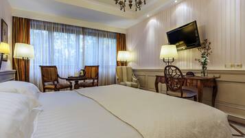 A hotel room with a large bed, two armchairs, a small table, a TV, and a chandelier.