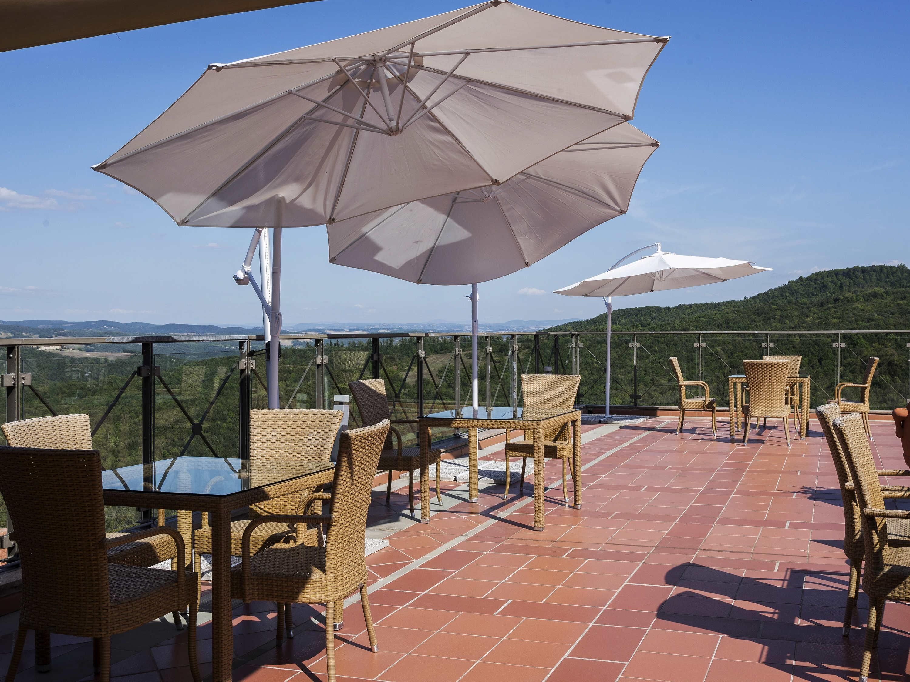 An outdoor patio with wicker chairs and tables, large umbrellas, and a scenic view of hills and mountains.