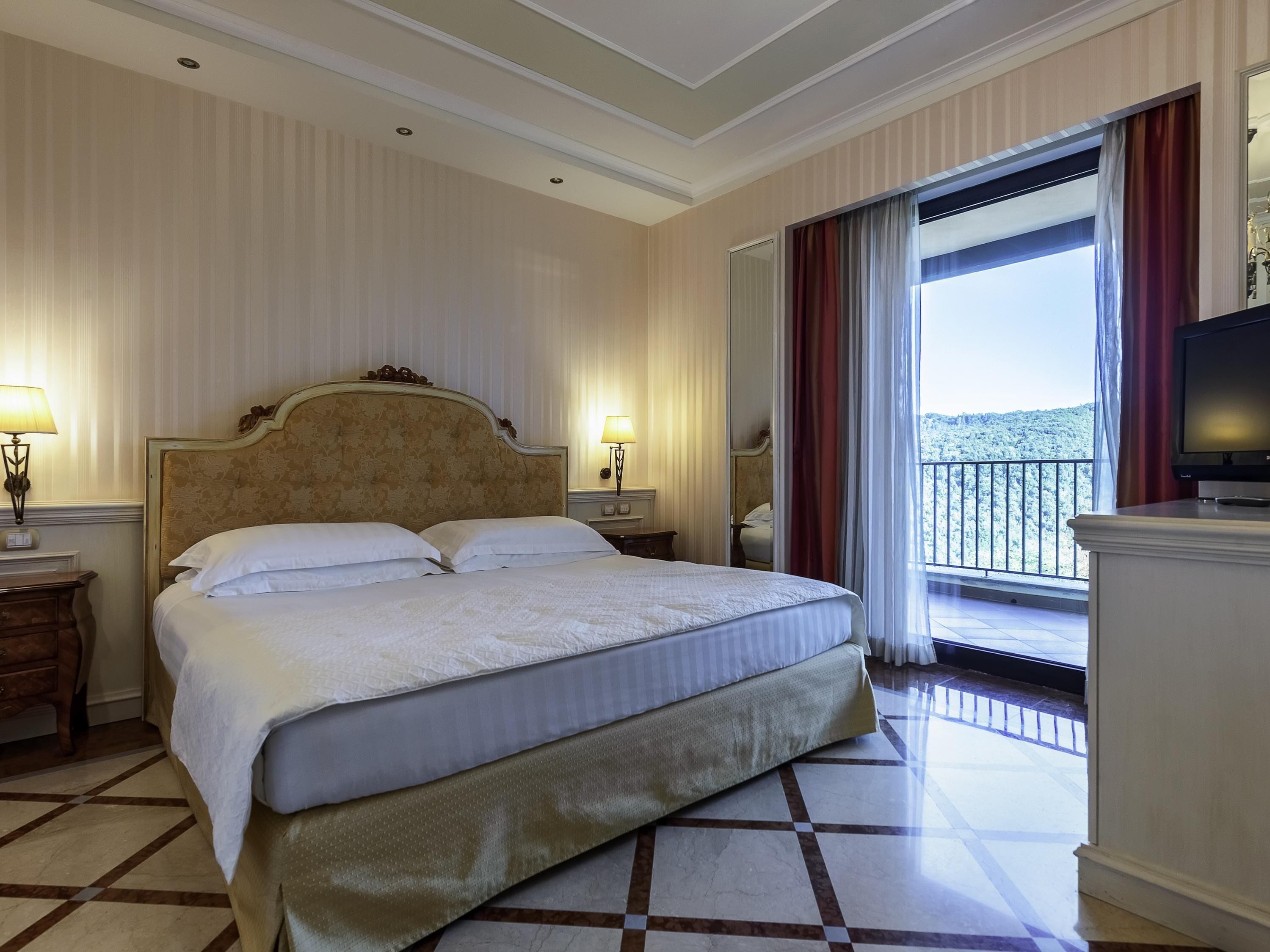 A hotel room with a large bed, two bedside tables, a TV, and a balcony with a view.