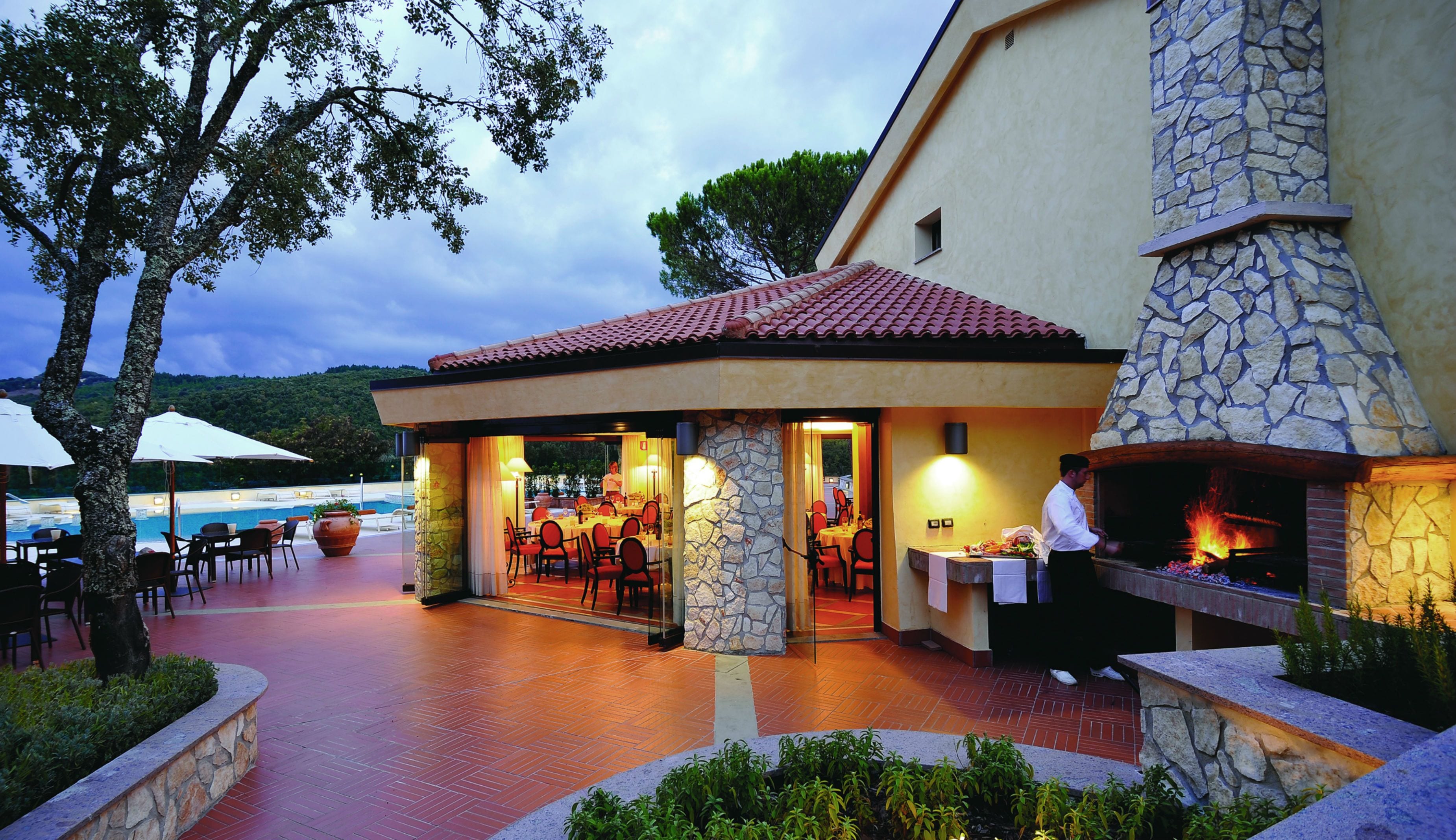An outdoor dining area with stone pillars, a stone fireplace, and a pool in the background.