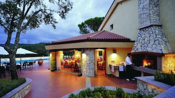 Un área de comedor al aire libre con pilares de piedra, chimenea de piedra y una piscina al fondo.