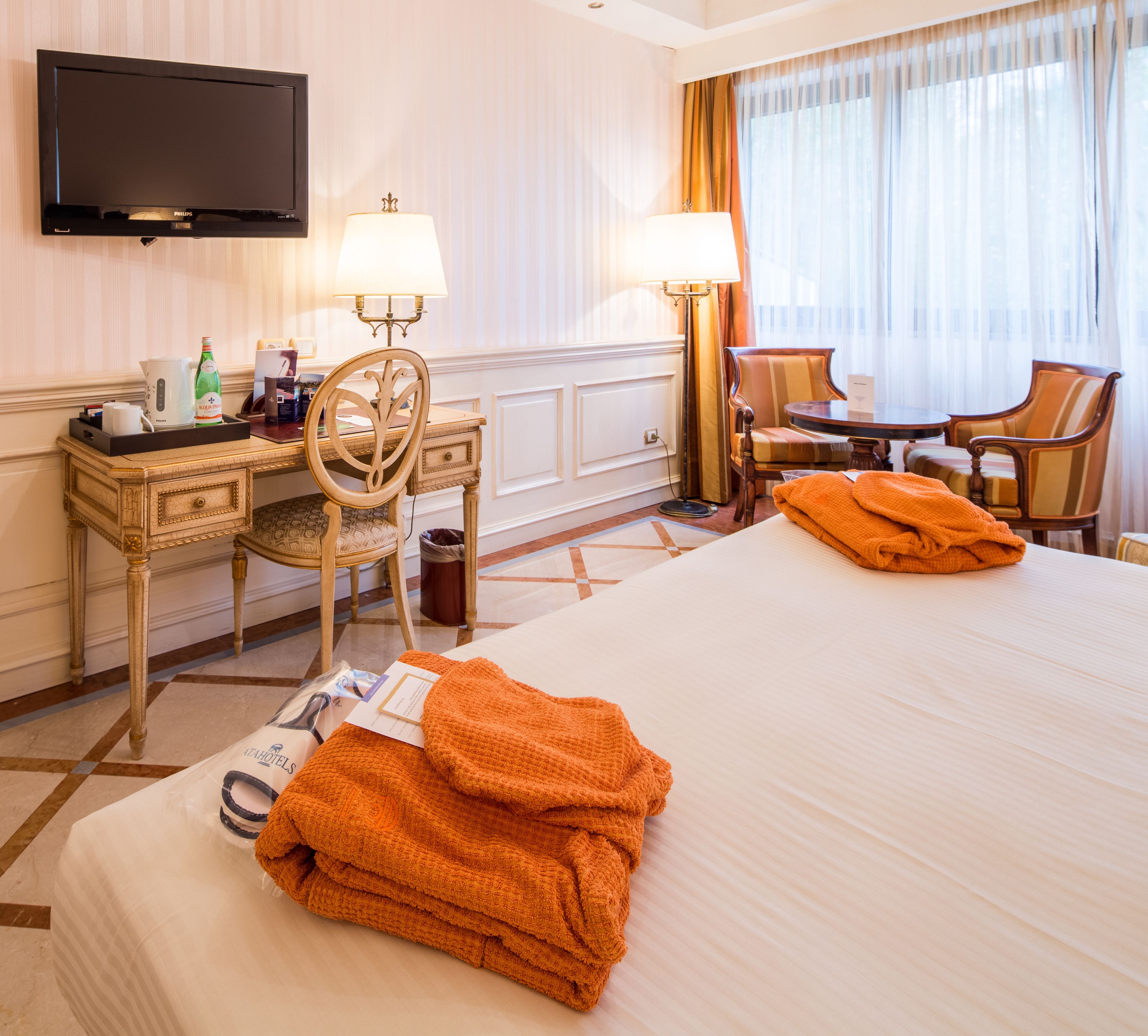 Camera d'albergo con un letto, due sedie, una scrivania con una lampada, una televisione e una finestra con tende.