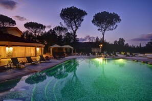 Piscina con sillones de descanso y una zona de estar cubierta, rodeada de árboles y un muro de piedra.