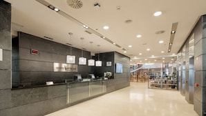 Un área de recepción de hotel moderna con un reloj digital que marca las 16:32, una mesa de recepción y una escalera que sube a los pisos superiores.