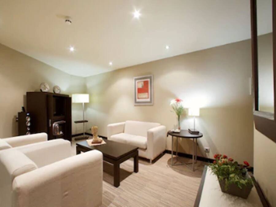 A modern living room with a sofa, armchairs, a coffee table, a side table with a lamp, a small cabinet, and a potted plant.
