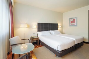 Habitación de hotel con cama doble, mesita de noche con lámpara, una silla y una pequeña mesa de vidrio.