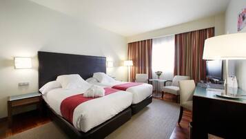 Quarto de hotel com duas camas, uma escrivaninha, uma cadeira, uma televisão e um abajur.
