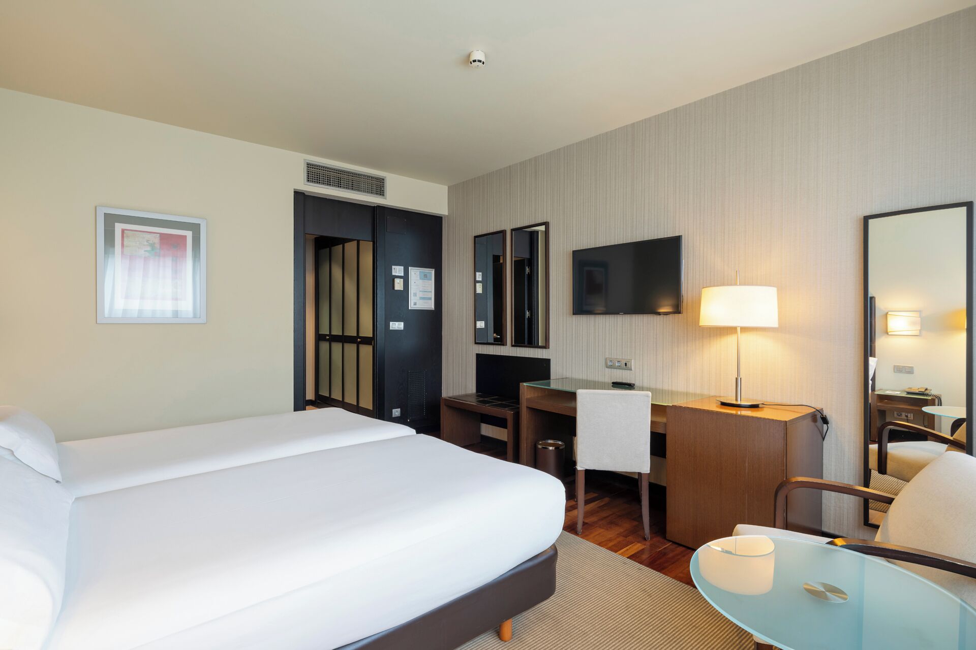 Una habitación de hotel con cama, escritorio, silla, televisor y una mesita de noche con lámpara.