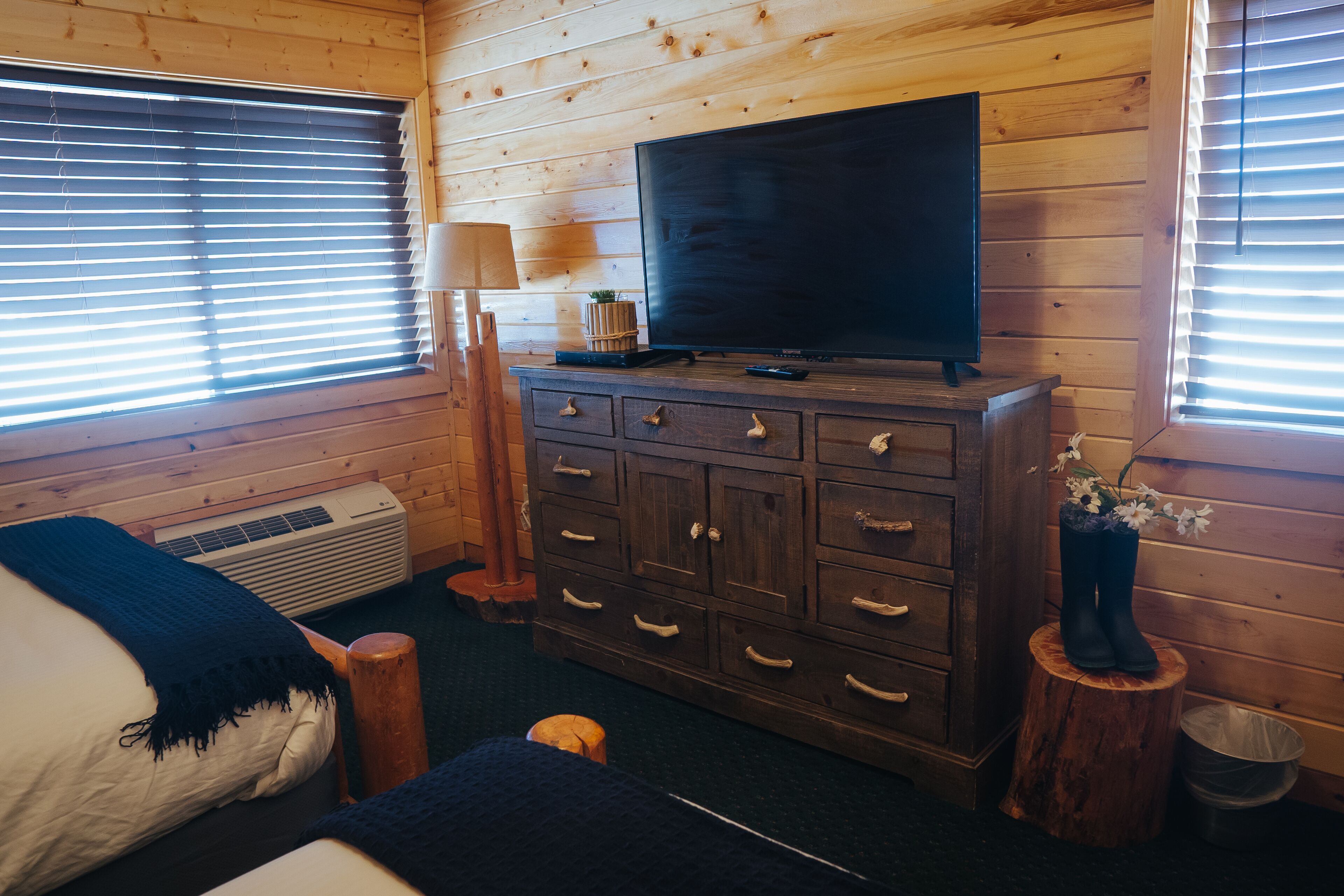 A wooden cabin room with a bed, dresser, TV, and windows.