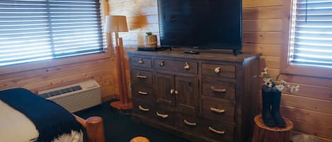 A wooden cabin room with a bed, dresser, TV, and windows.
