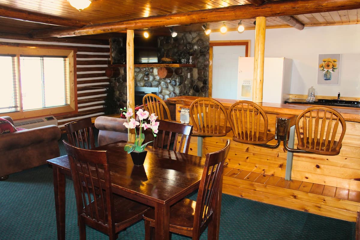 A wooden dining table with chairs, a stone fireplace, and a kitchen area with a refrigerator and sink.