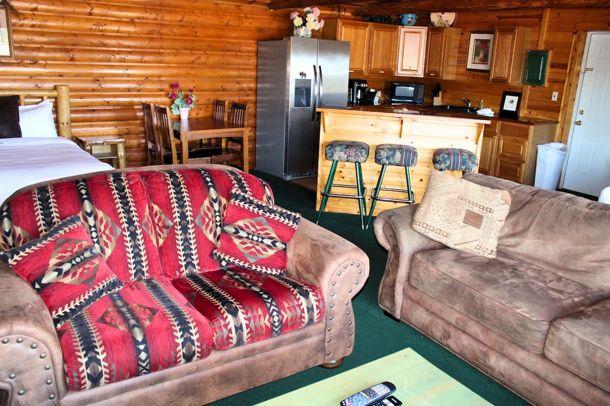 A log cabin interior with a bed, two sofas, a kitchen area, and a dining table.