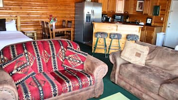 A log cabin interior with a bed, two sofas, a kitchen area, and a dining table.