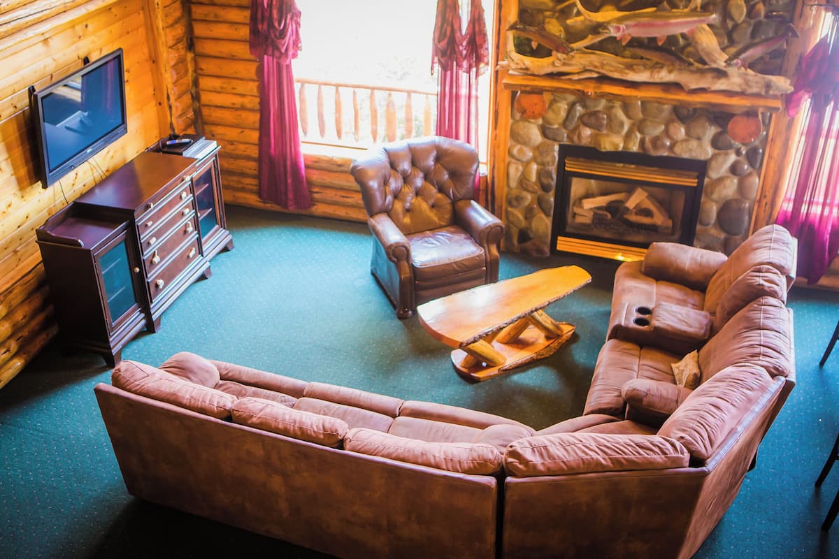 A log cabin interior with a leather sofa, a wooden coffee table, a stone fireplace, and a television on a wooden cabinet.