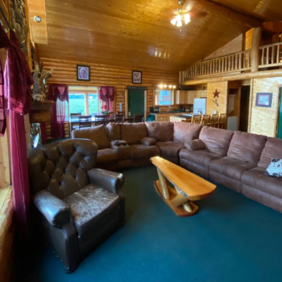 Een blokhut interieur met een grote leren bank, een houten salontafel, en een eethoek met een hoge plafond en houten balken.