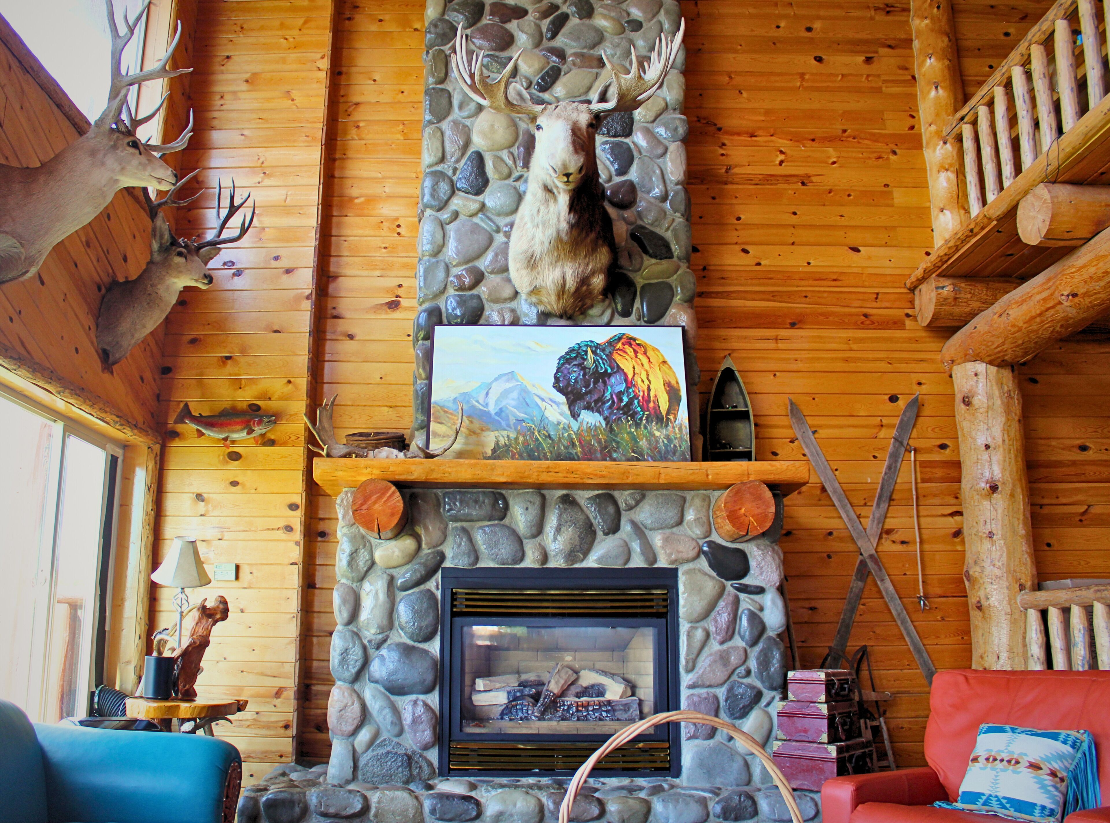 A log cabin interior with a stone fireplace, wooden walls, and a mounted deer head.